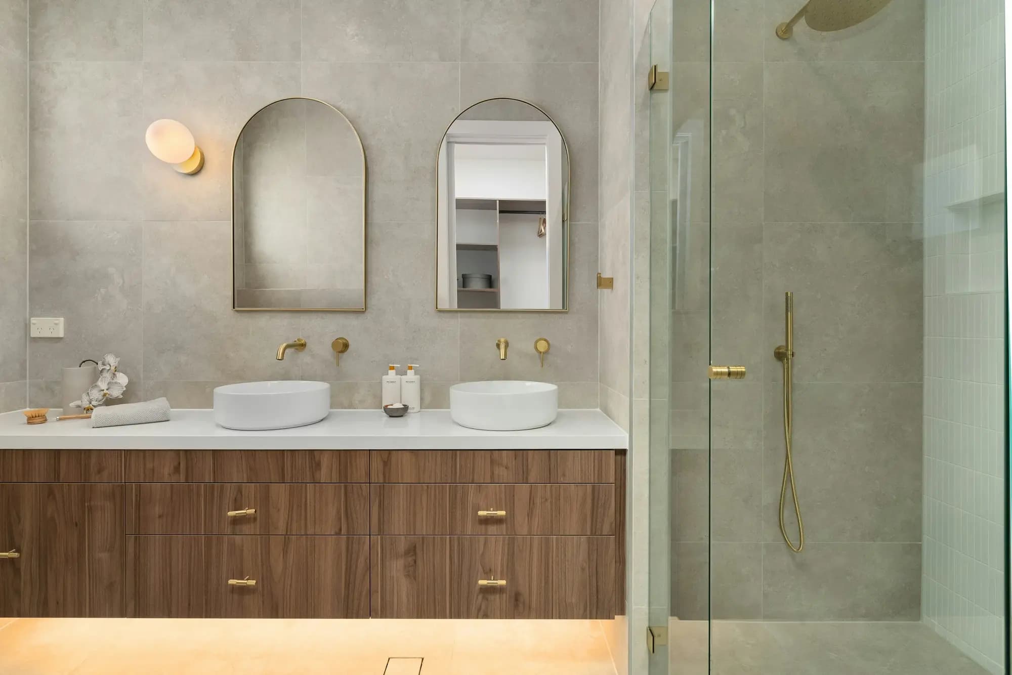 Double timber vanity with twin basins, arched mirrors, brushed brass tapware and a frameless shower screen