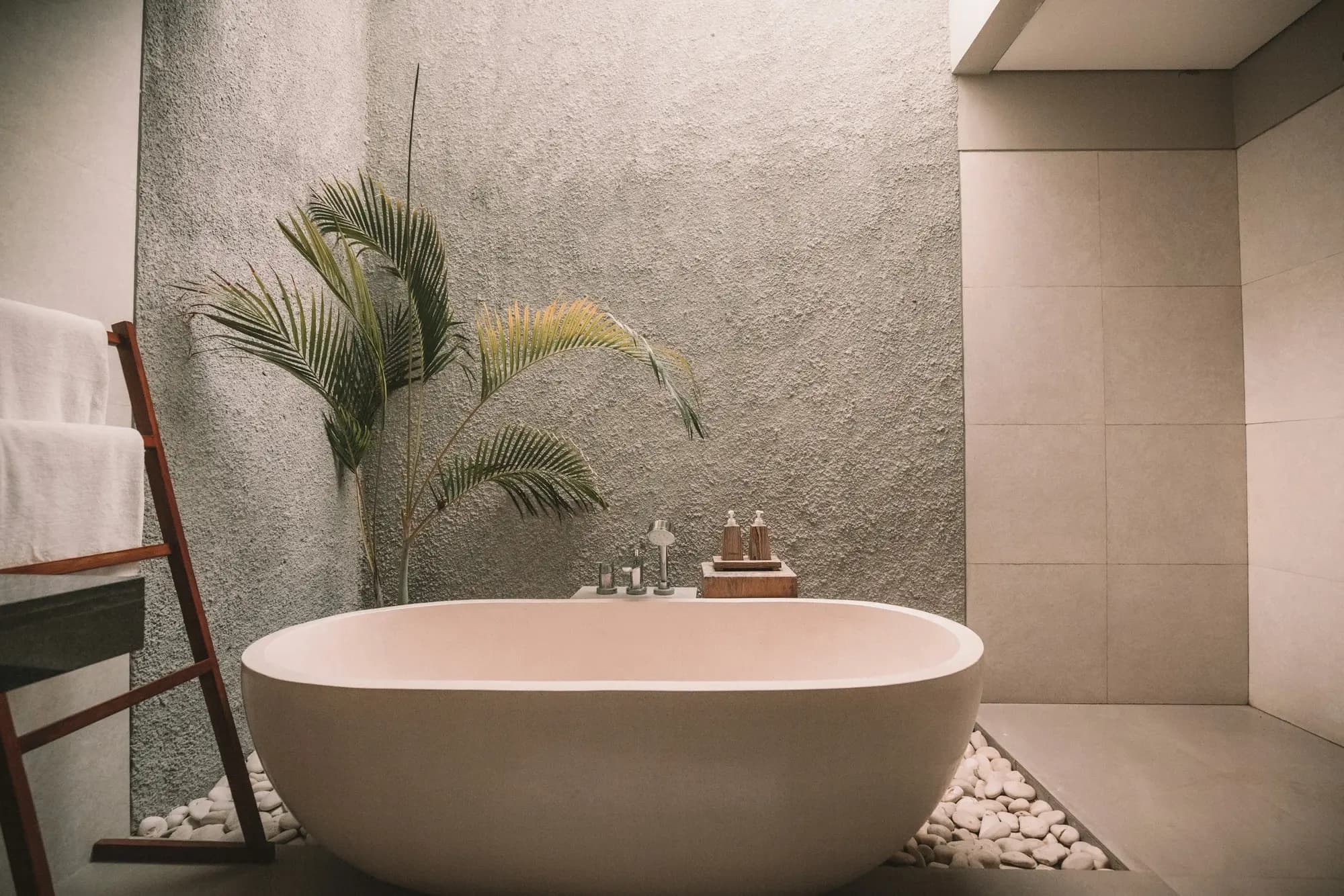 Freestanding stone bath on a pebble bed beside a rendered feature wall and a palm