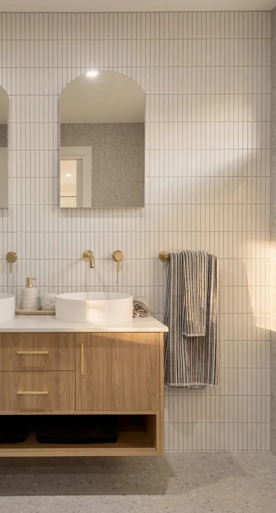 Timber wall-hung vanity with a vessel basin, brushed brass tapware, arched mirror and vertical finger tiles